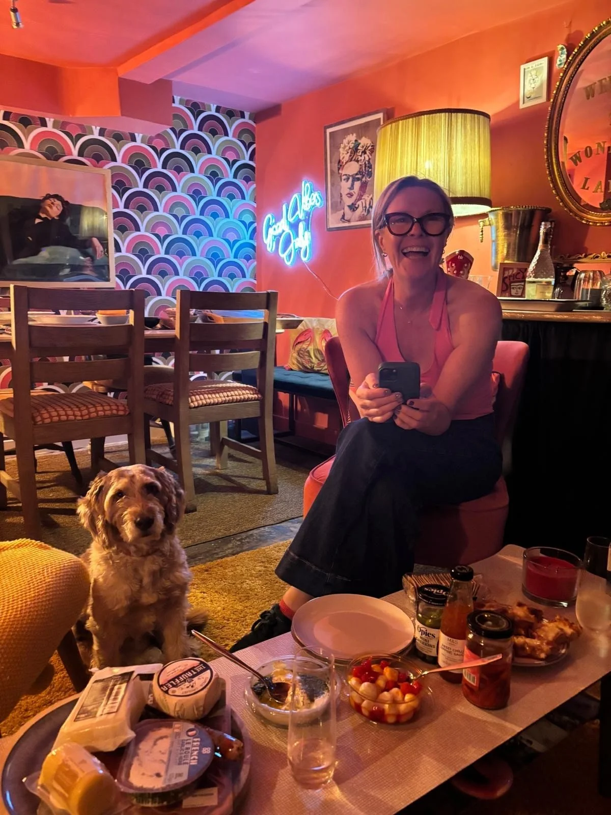 A woman with glasses smiling and holding a phone, sitting in a cozy, colorful indoor setting with a dog at her feet. The room features a vibrant patterned wall, a neon sign, various decorations, a dining table with food, and warm lighting.