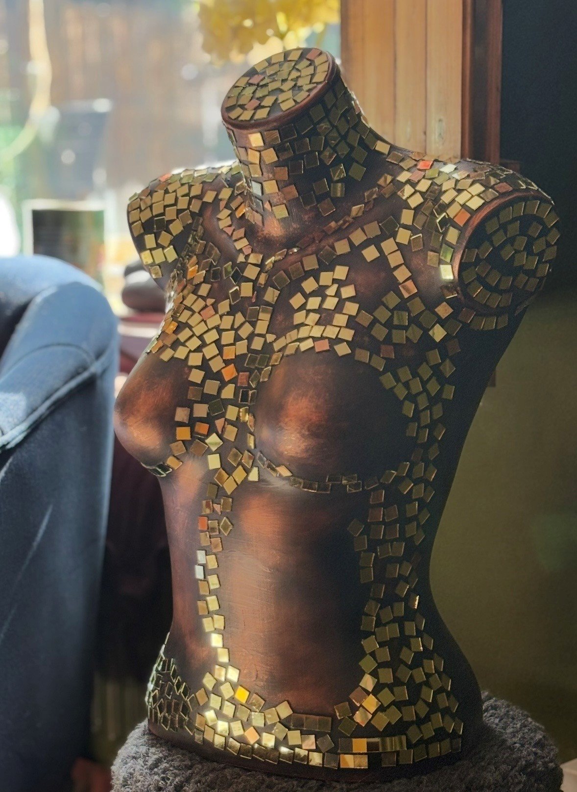 An artistic sculpture of a human torso decorated with small, shiny, gold-colored mosaic tiles, mounted on a stand.