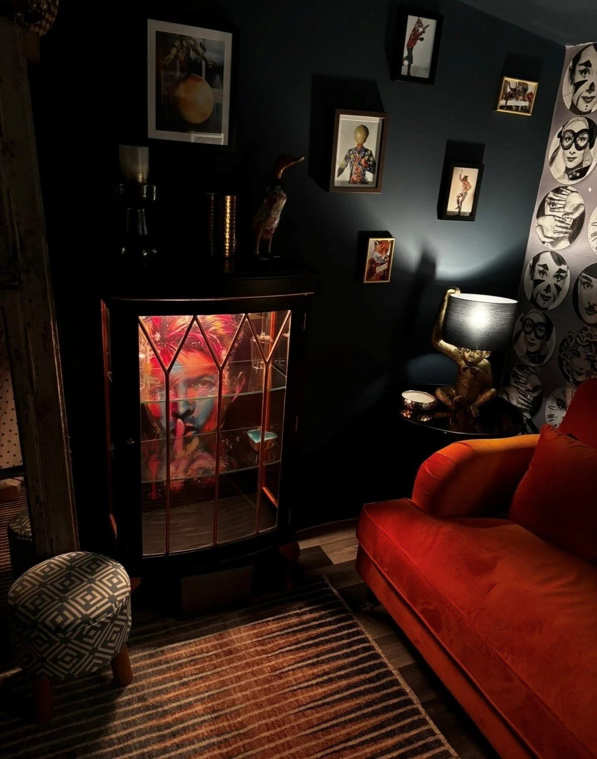 A cozy living room corner decorated with framed artwork on dark walls, a black cabinet with a colorful portrait inside, a black side table with a unique gold sculpture lamp, and an orange velvet armchair, all illuminated by a table lamp and accent li