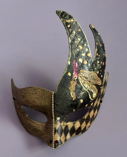 A decorative mask with a crescent moon shape, half painted in gold with a checkered pattern and the other half decorated with a large butterfly design.