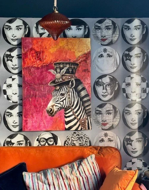 Interior room with a red, orange, and yellow abstract background painting of a zebra with a hat, surrounded by black and white face images on the wall, decorative hanging light, and orange sofa with striped and orange pillows.