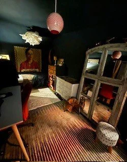 Decorative room with a colorful portrait on the wall, a round pink paper lantern hanging from the ceiling, and various vintage furniture pieces including a mirror and a chair.
