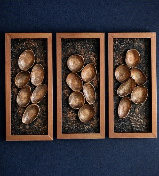 Three wooden framed shadow boxes mounted on a dark wall, each containing oval metallic bowls and a dark, textured background.