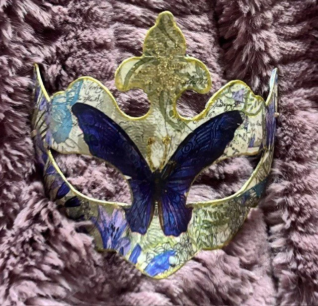 Decorative mask with a large blue butterfly design, placed on a soft, textured purple surface.