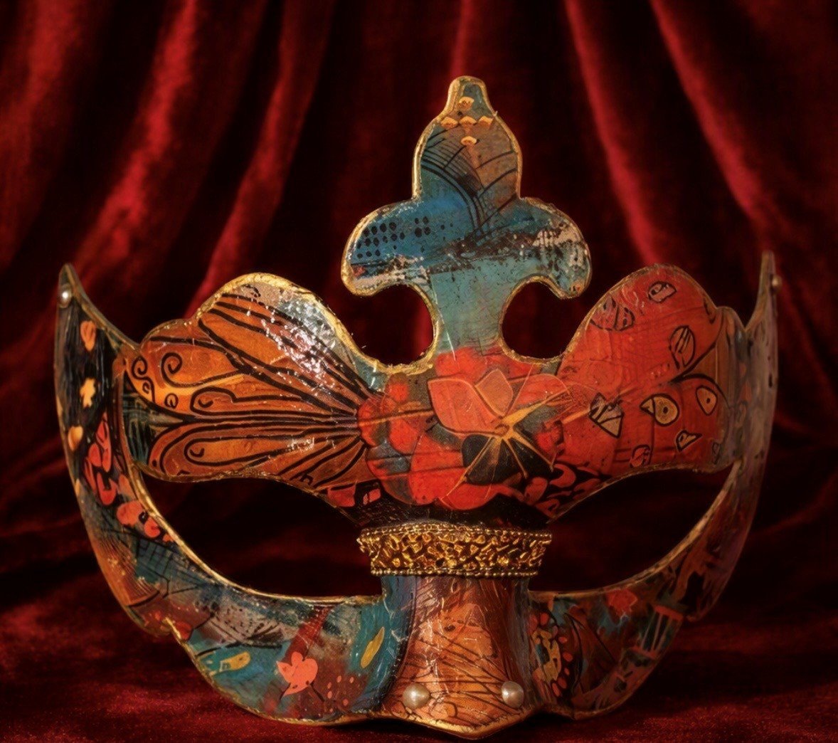 A decorative mask with intricate patterns and bright colors, resembling a crown with a butterfly and floral motifs, set against a dark red velvet background.