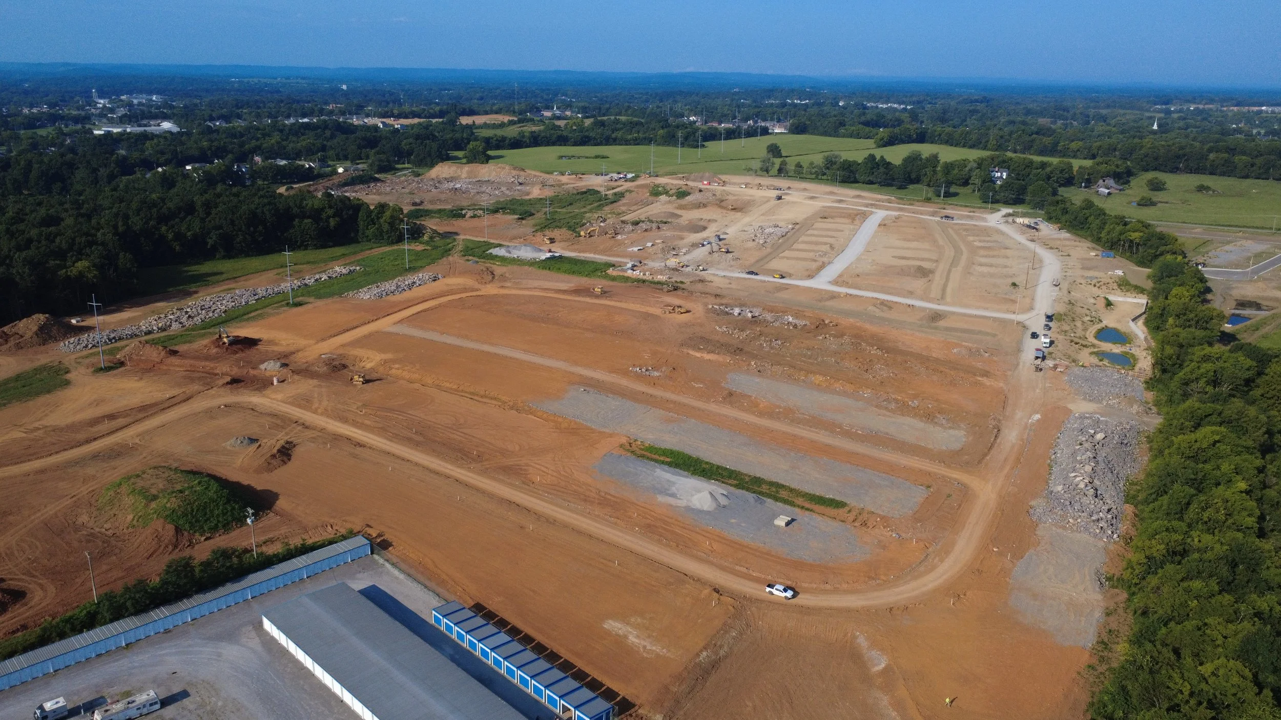 Aerial view of a construction site with dirt roads, excavators, and construction vehicles surrounded by green trees and fields.