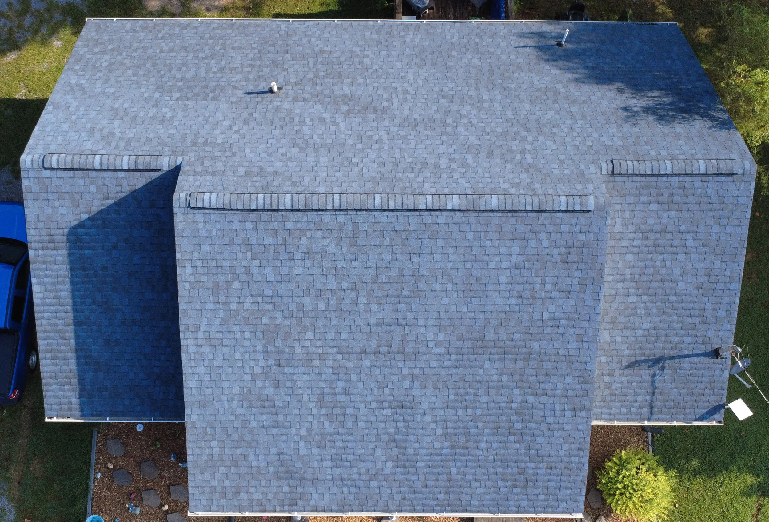 An aerial view of a house with a gray shingled roof, featuring vents and outdoor piping, surrounded by trees, grass, and a blue car parked on the driveway.