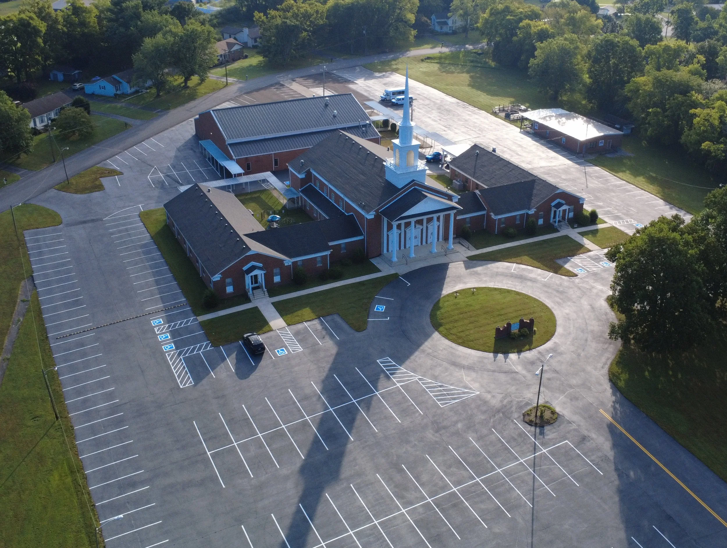 A church with a white steeple and brick walls, surrounded by a large parking lot with marked parking spaces and trees.