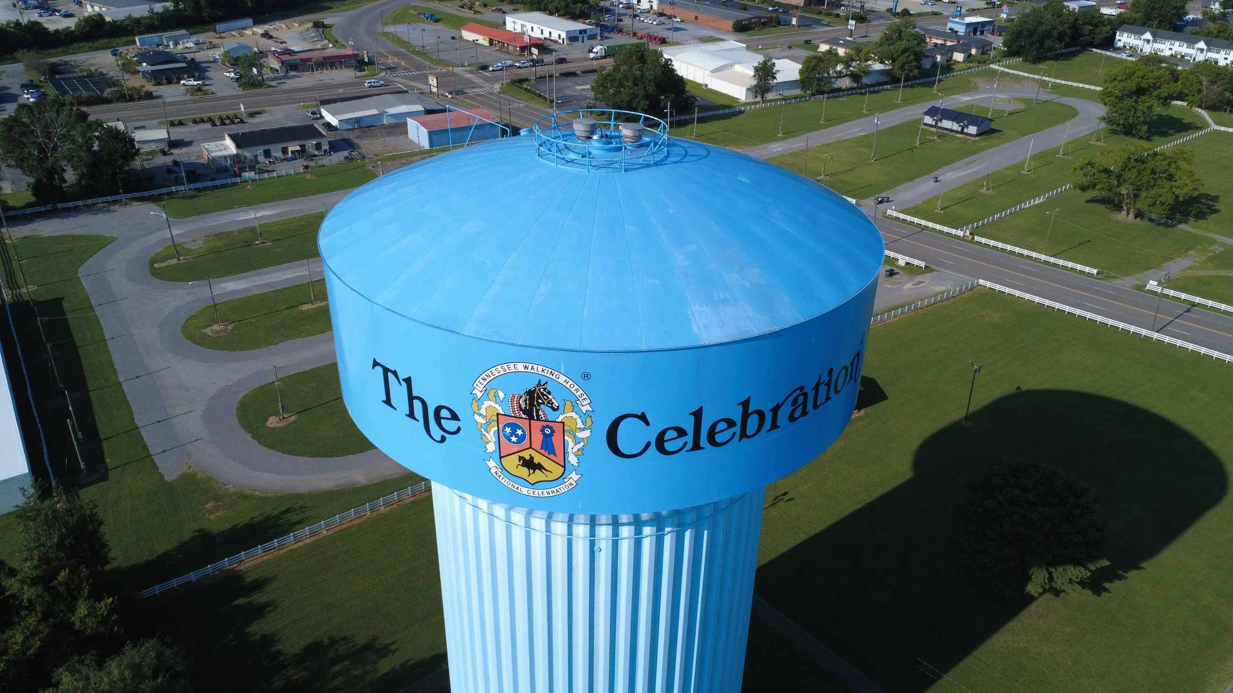 Aerial view of a large blue water tower with the words 'The Celebration' and a crest featuring a horse and various symbols. Surrounding the tower are roads, parking lots, and grassy areas with trees.