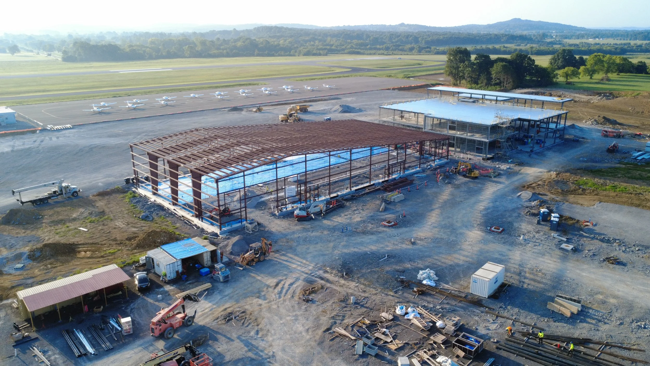 Aerial view of a construction site near an airport with planes parked on the tarmac and a partially built structure with construction equipment and workers.
