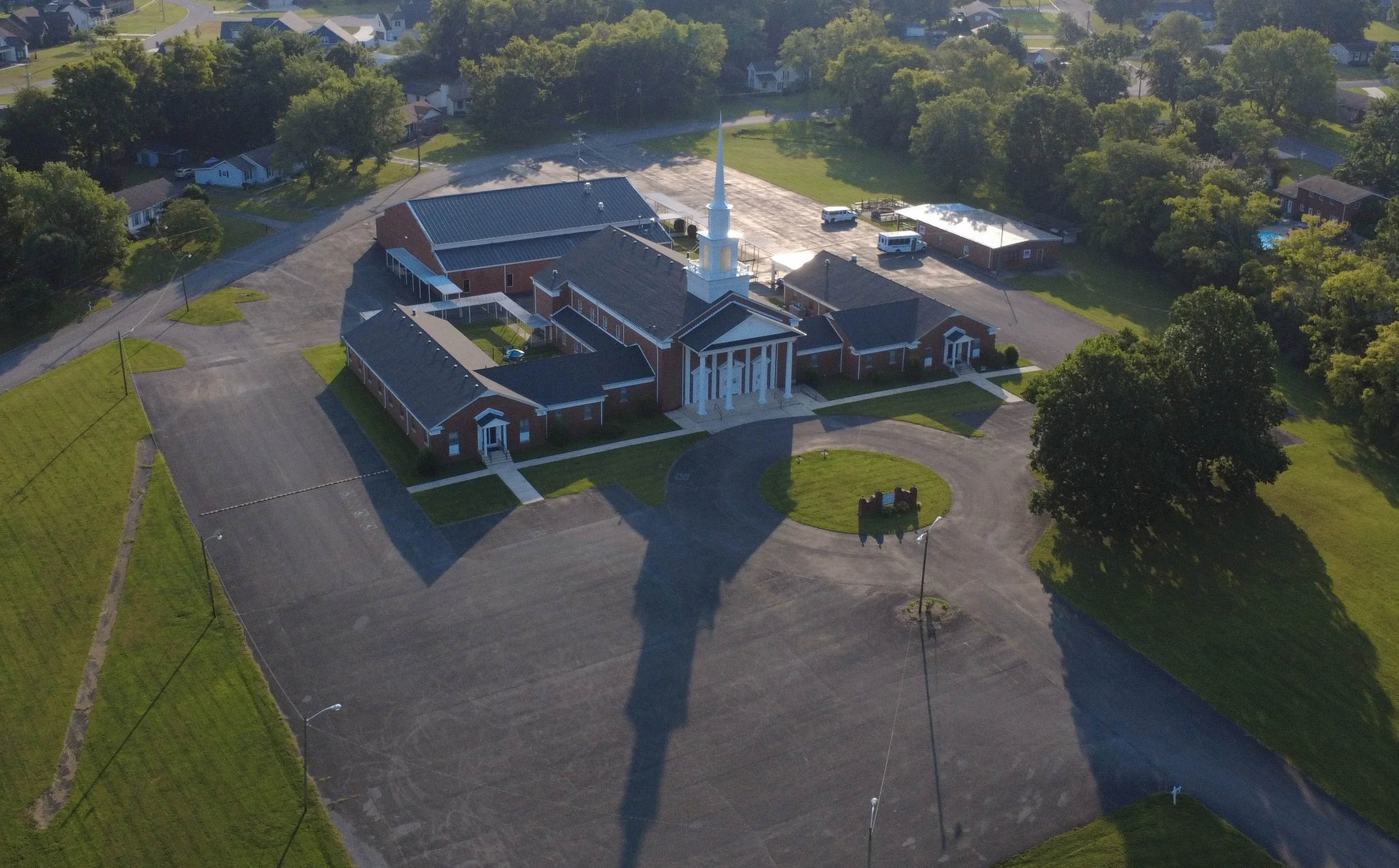 Aerial view of a church with a prominent steeple, surrounded by a parking lot, green lawns, and residential neighborhood with trees and houses.