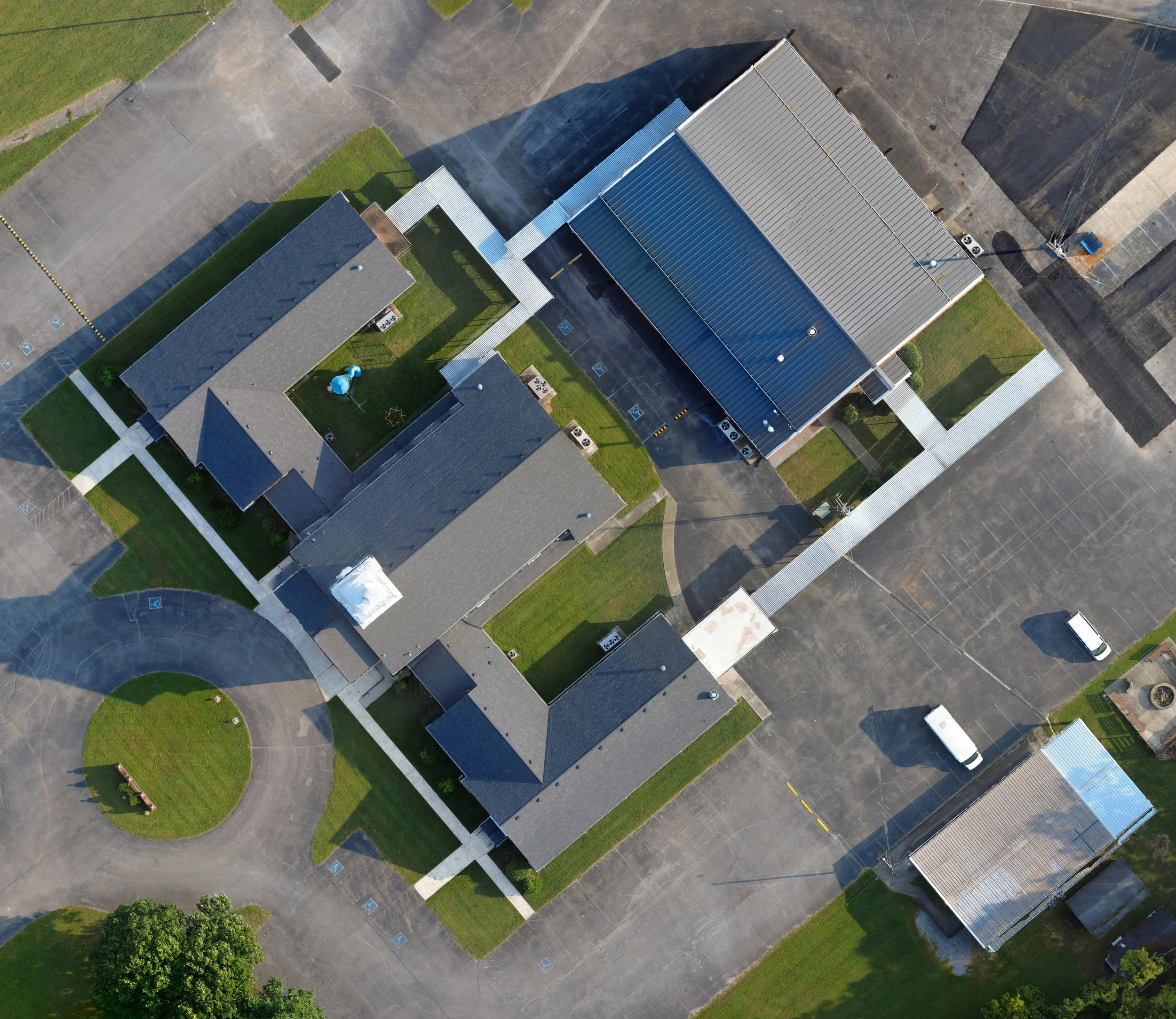 Aerial view of a building with a metal roof, parking lot with cars, grassy areas, and sidewalks.