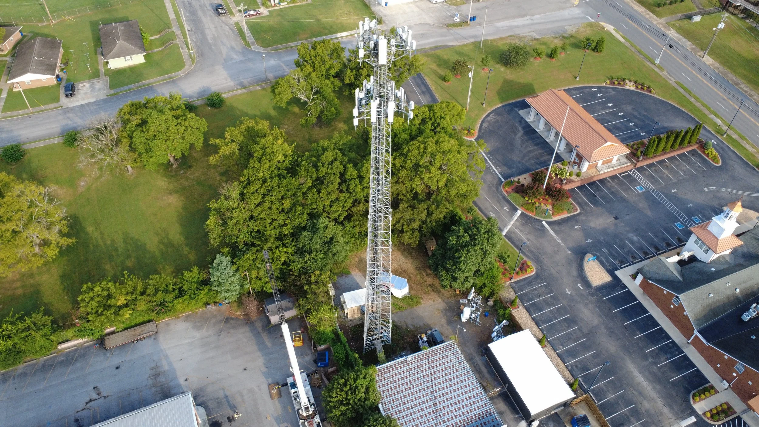 An aerial view of a suburban area with trees, a communication tower, a parking lot, and buildings.