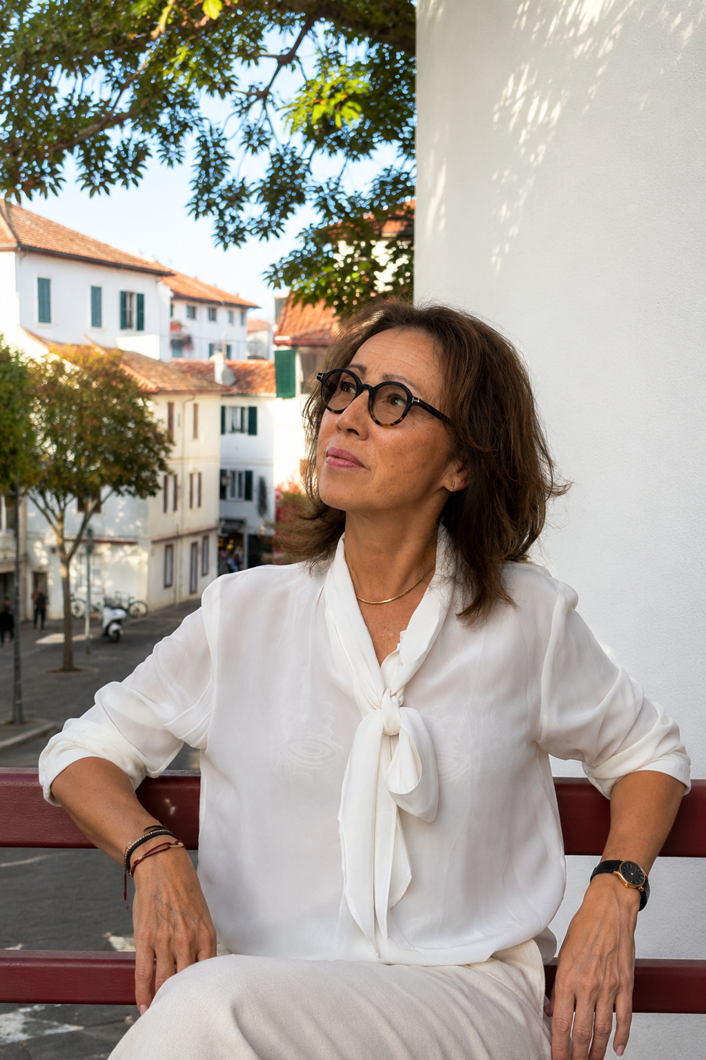 Une femme âgée avec des cheveux bruns et frisés, portant des lunettes rondes, une chemise blanche avec un nœud à l'encolure, assise sur un banc en bois rouge dehors, avec des bâtiments blancs et des arbres en arrière-plan.