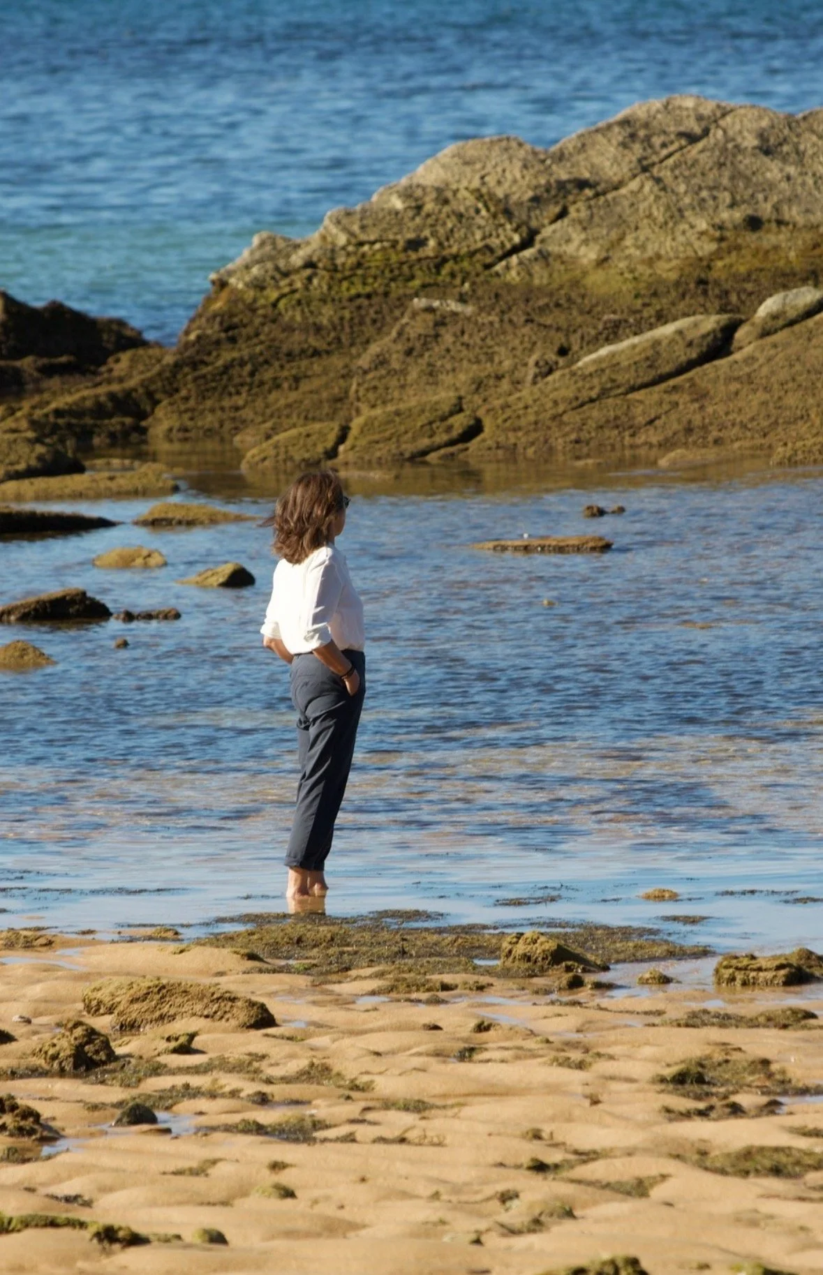 Une femme debout dans l'eau sur une plage rocheuse, regardant vers la mer.