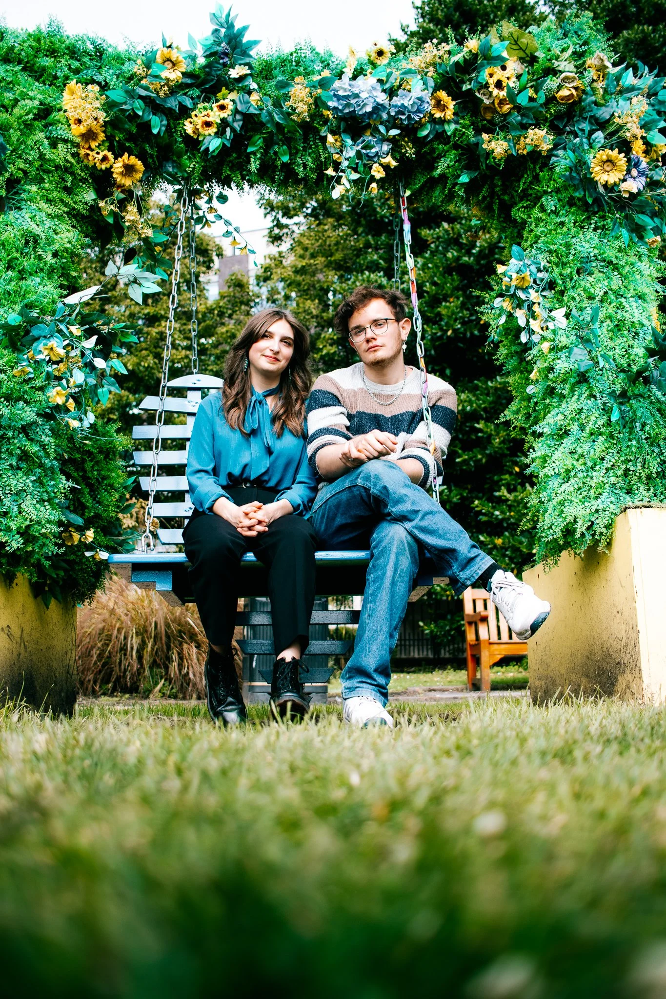 Young man and woman sitting on a swing surrounded by lush green foliage and flowers in an outdoor garden setting.