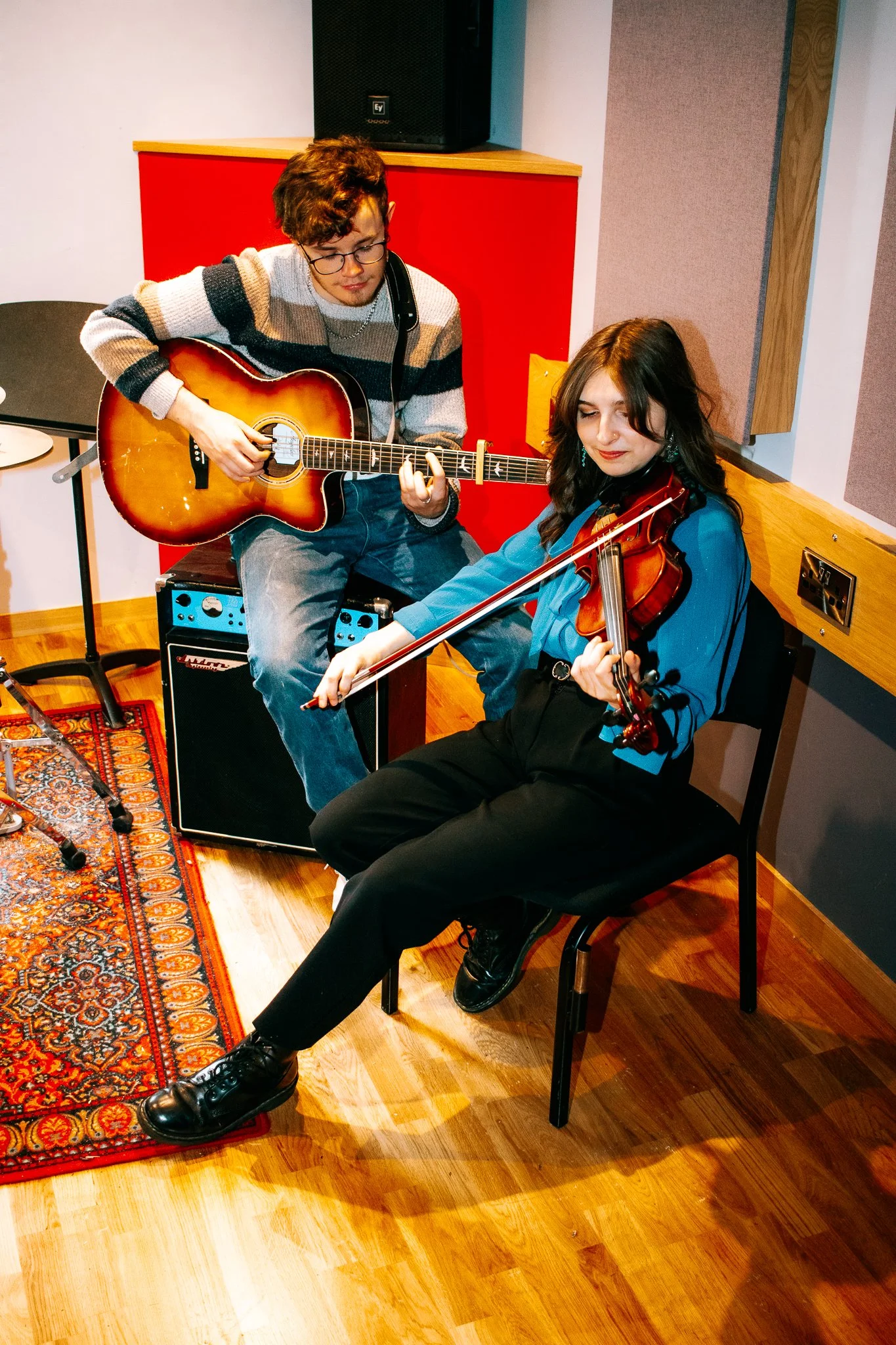 Young man with glasses playing an acoustic guitar, young woman with long dark hair playing a violin, in a music studio with wood flooring, upholstery, and an amplifier.