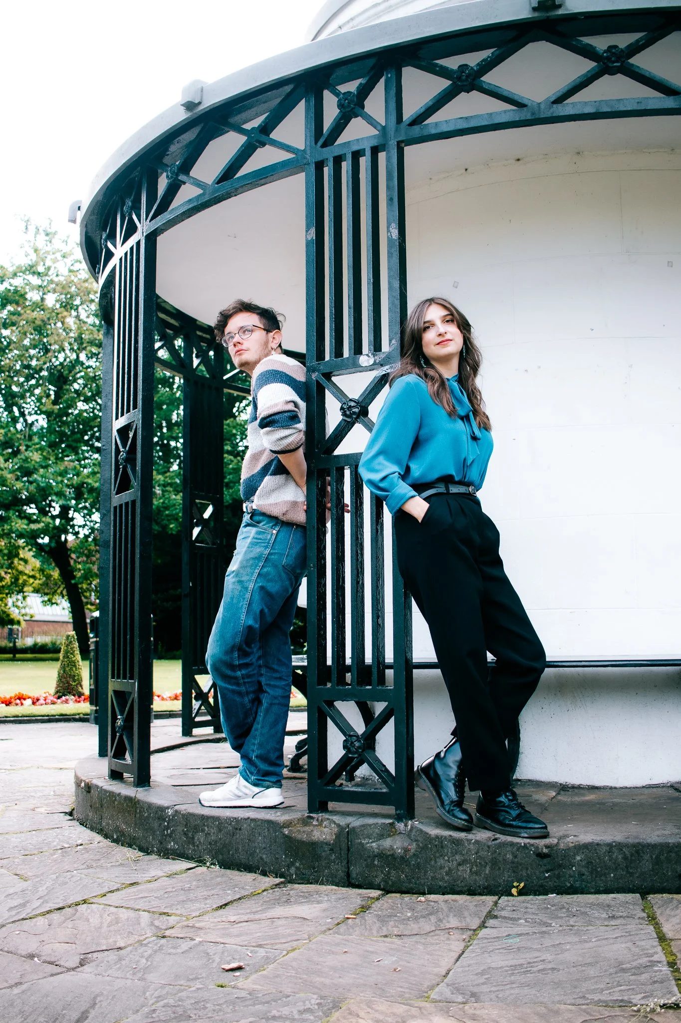 A man and woman leaning against a black metal gate around a white building, with greenery and flowers in the background.