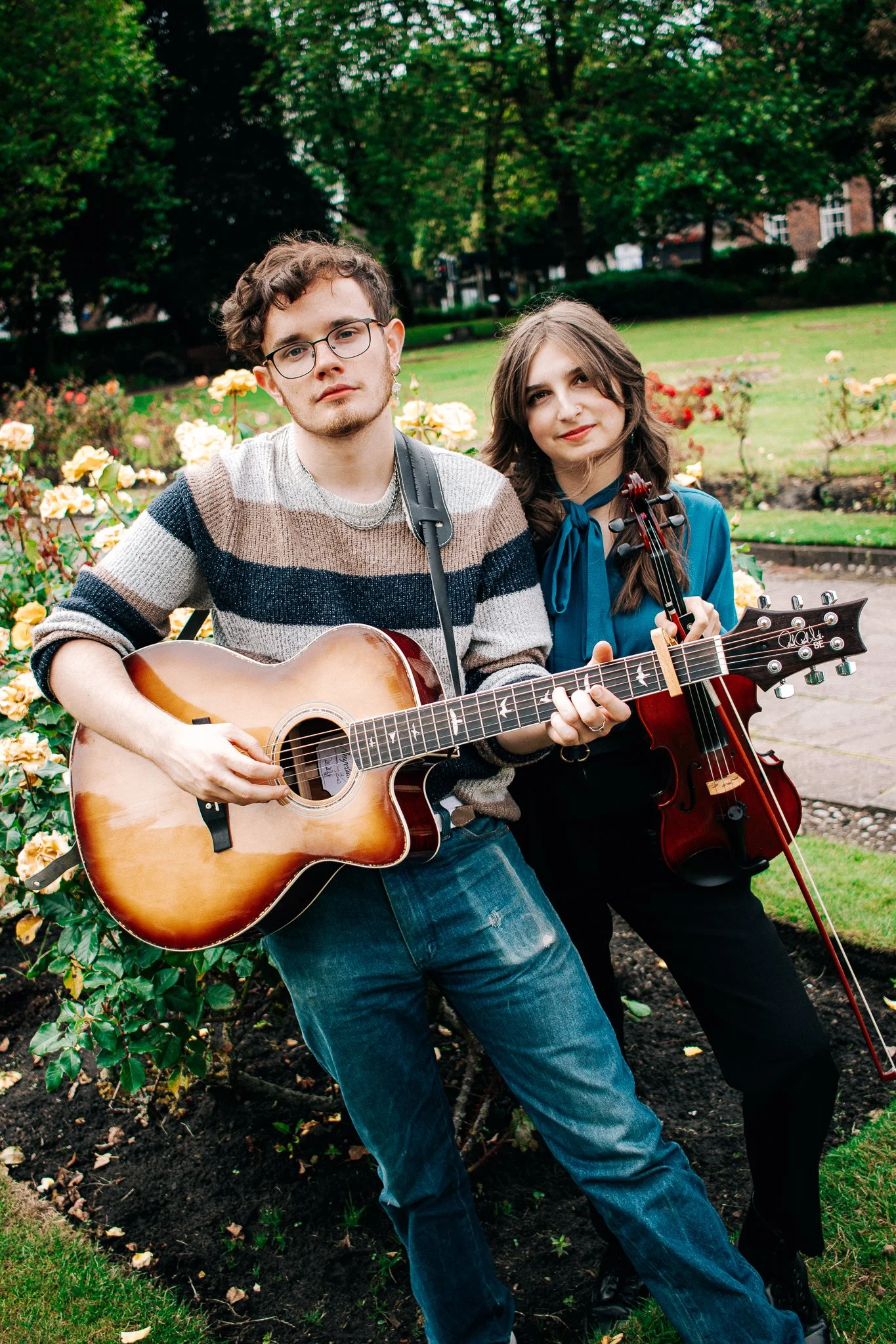 A young man and woman outdoors in a garden, playing musical instruments. The man is playing an acoustic guitar, wearing glasses and a striped sweater. The woman is holding a violin, with long brown hair and a blue top, smiling slightly. The background features green trees, a lawn, and blooming flowers.