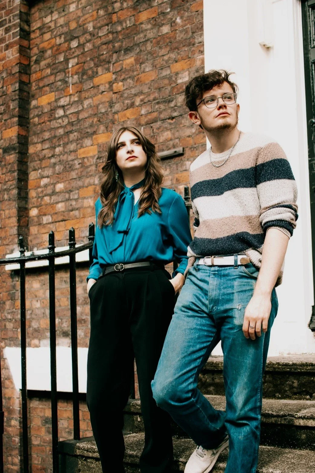 Two young adults, a woman in a teal blouse and formal black pants and a man in a striped sweater and jeans, standing outside on a staircase with a brick wall background.