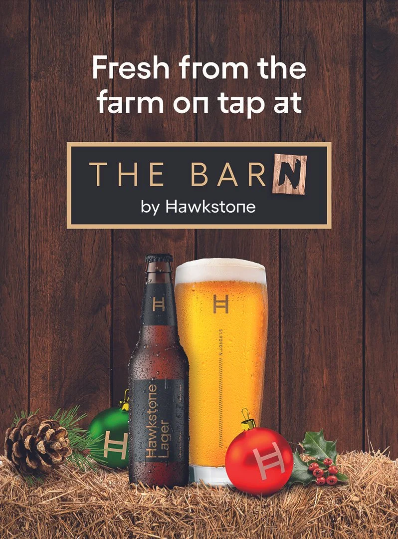 A promotional image for Hawkstone beer featuring a bottle and a glass of beer on a bed of straw, with Christmas ornaments, holly leaves, and pinecones, set against a wooden background.