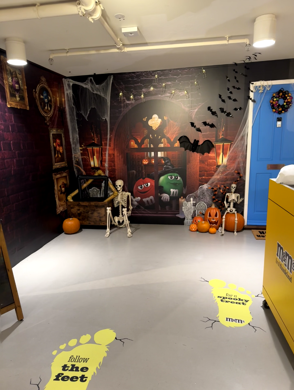 Halloween-themed indoor display with a spooky backdrop of a haunted house, pumpkins, skeletons, bats, spider webs, and M&M characters in costumes, decorated for Halloween.