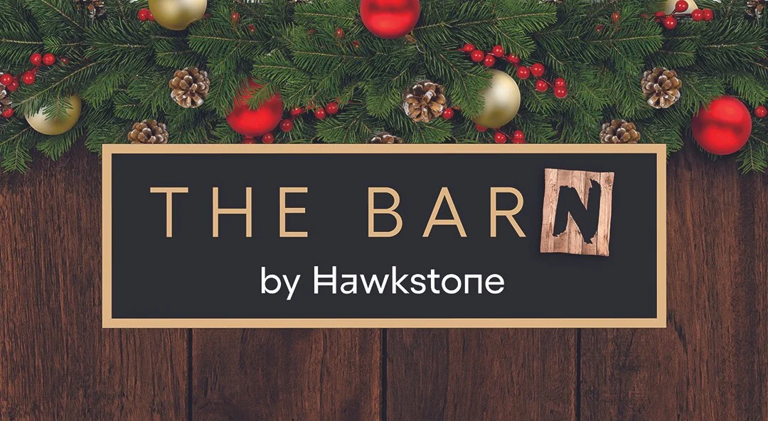 Decorative image with green pine branches, red and gold ornaments, pinecones, and red berries, with a sign reading 'The Barn by Hawkstone' placed over a wooden background.