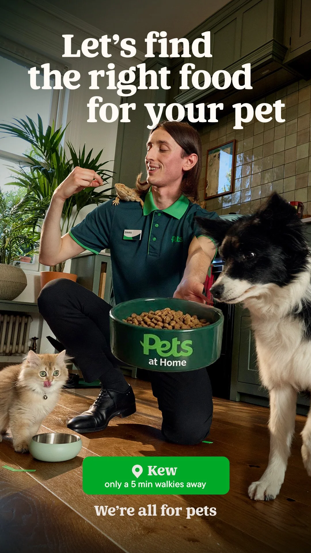 A man with long hair, wearing a Pets at Home uniform, feeding his pets in a cozy kitchen. There is a cat licking its nose, a Border Collie dog sniffing the food, and a lizard on the man's shoulder. A large green pet food bowl is in the center, and a 