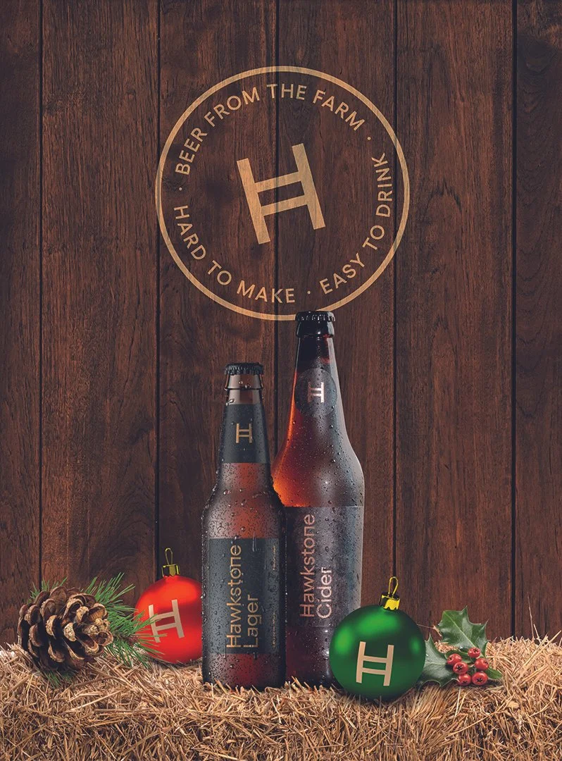 Two bottles of Hawkwstone cider and two decorated Christmas ornaments, one red and one green, arranged on a bed of straw with pinecones and holly, against a wooden background with a circular logo reading 'H' and 'Beer from the farm, hard to make, eas