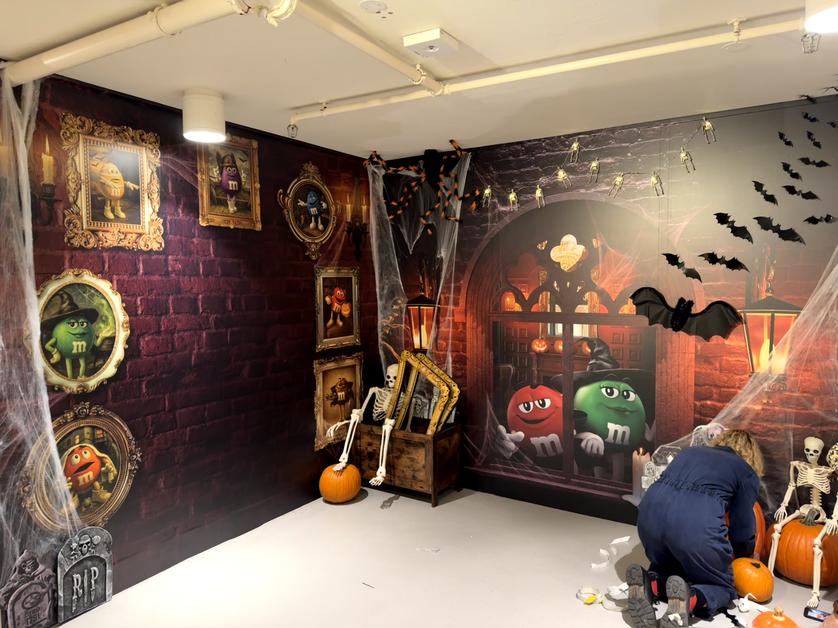 Haunted Halloween scene with M&M characters, framed portraits, skeleton, pumpkins, spider webs, bats, skulls, and spooky decor.