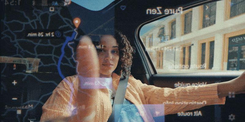 Young woman with curly hair sitting in a car with a GPS navigation screen in the windshield showing a map and route, while driving through a city street with buildings in the background.