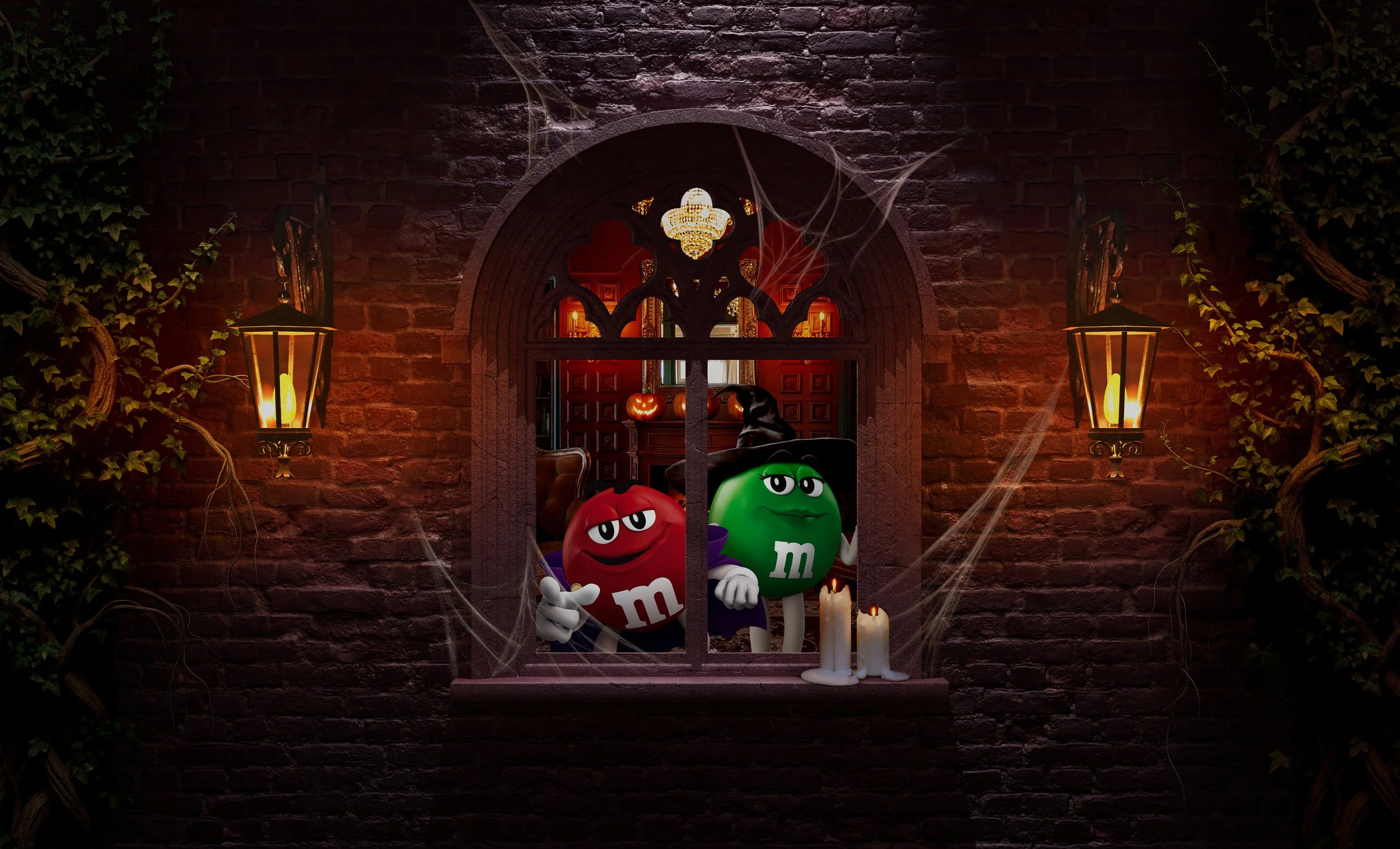 Two animated M&M characters, a red and a green one, dressed as witches, standing behind a window with cobwebs. The scene is decorated with candles and Halloween pumpkins, set against a brick wall background illuminated by lanterns.