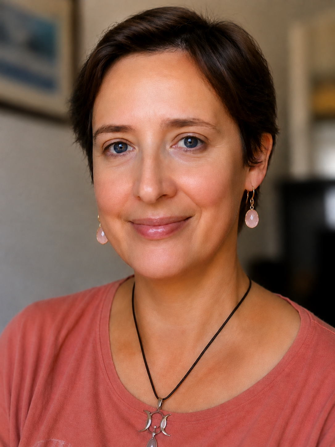 Une femme souriante aux cheveux courts, portant des boucles d'oreilles en pierre rose, un collier avec un pendentif en forme de lune, et un vêtement de couleur corail, dans un cadre intérieur.