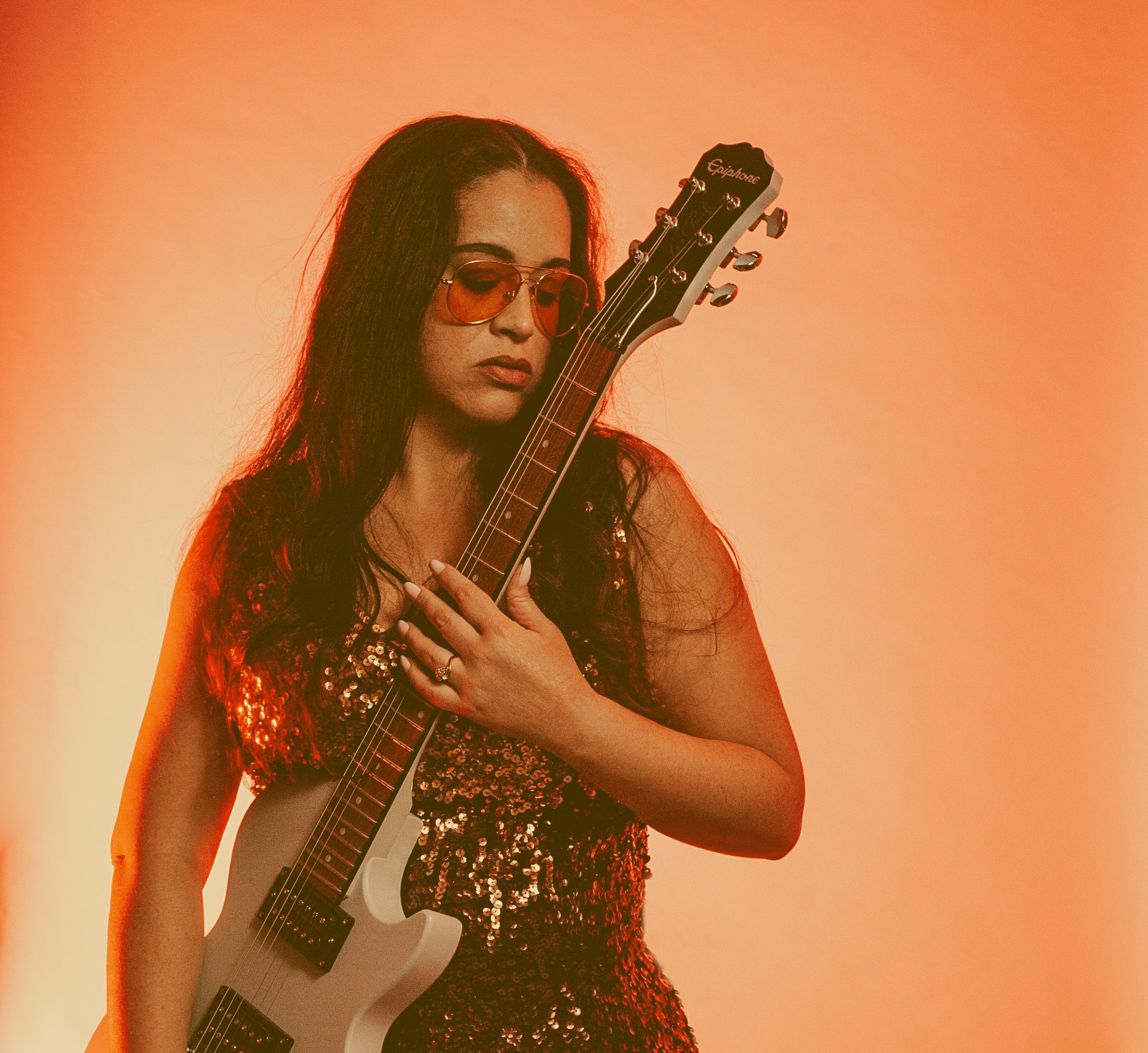 A woman with long dark hair wearing sunglasses and a sequined dress, holding an electric guitar against a warm orange background.