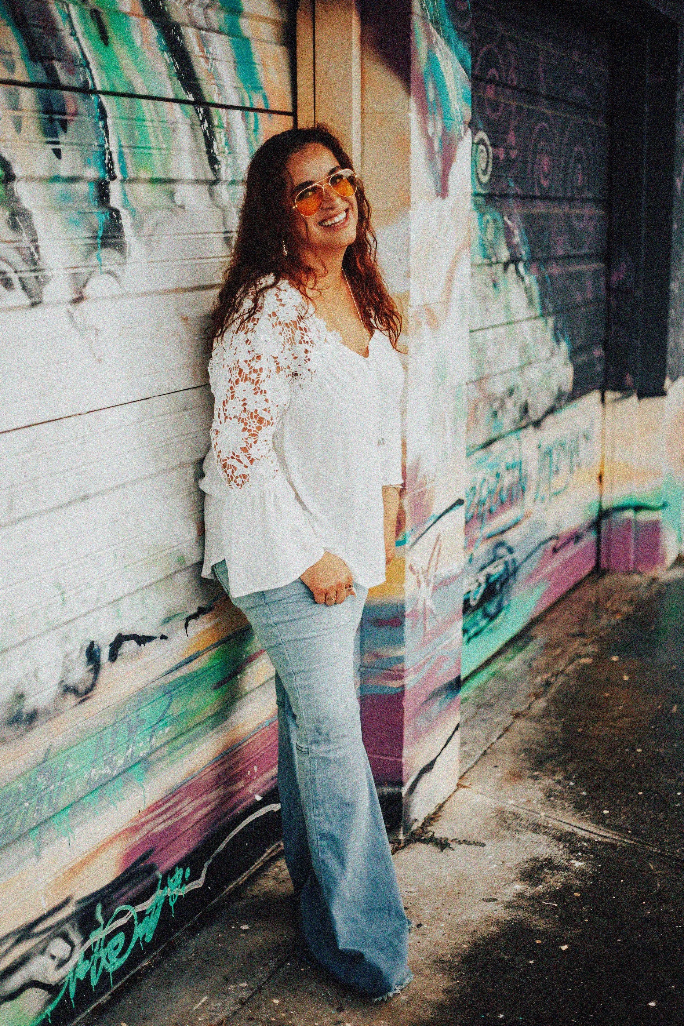 A woman with curly hair and orange sunglasses smiling and leaning against a colorful graffiti-covered wall.