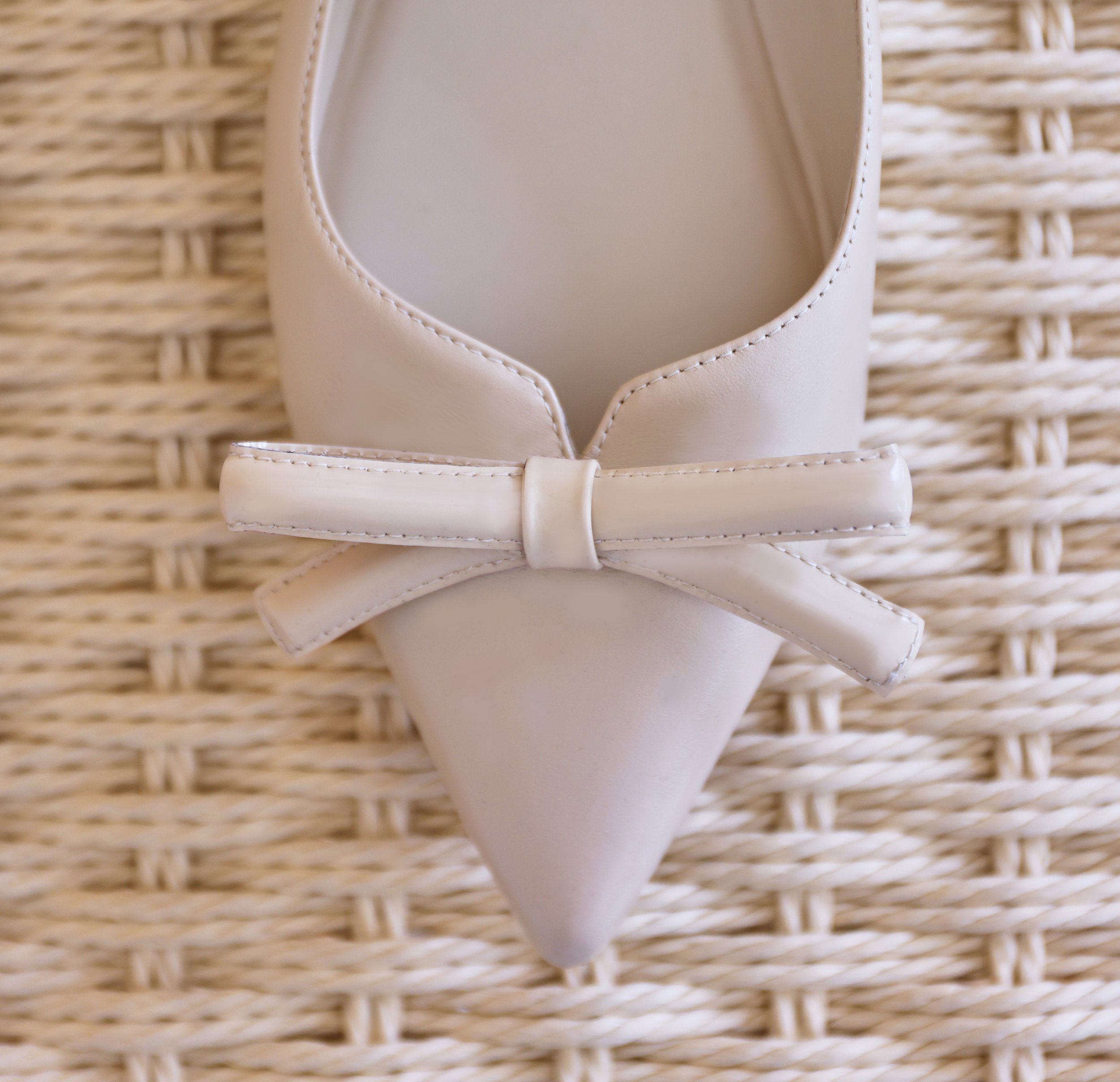 Close-up of a white leather sandal with a bow on a woven basket surface.