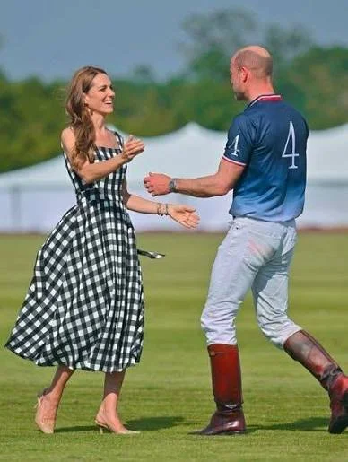 A woman in a black and white checkered dress and high heels is meeting a man in a blue sports jersey and riding boots on a grassy field, with tents and trees in the background.