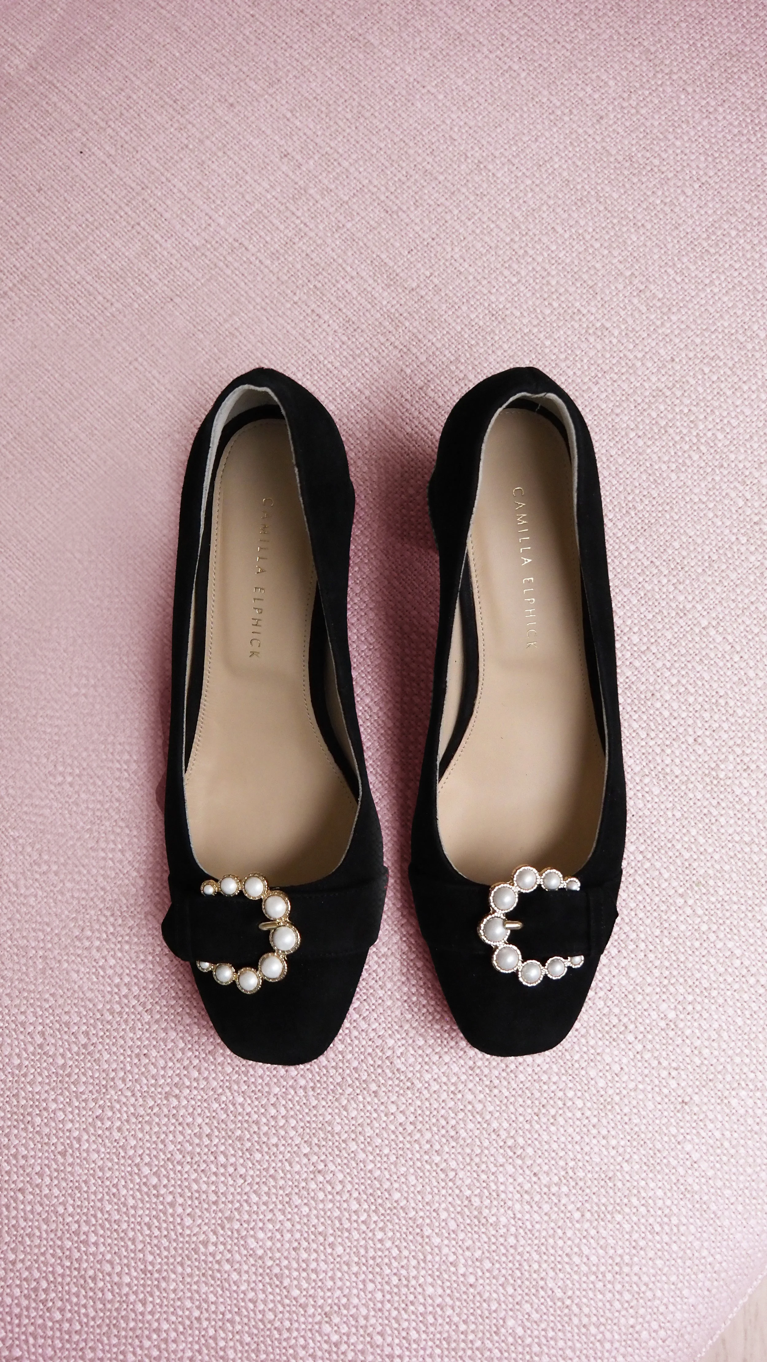 A pair of black pointed-toe flats with pearl and gold embellishments on the front, placed on a pink textured surface.
