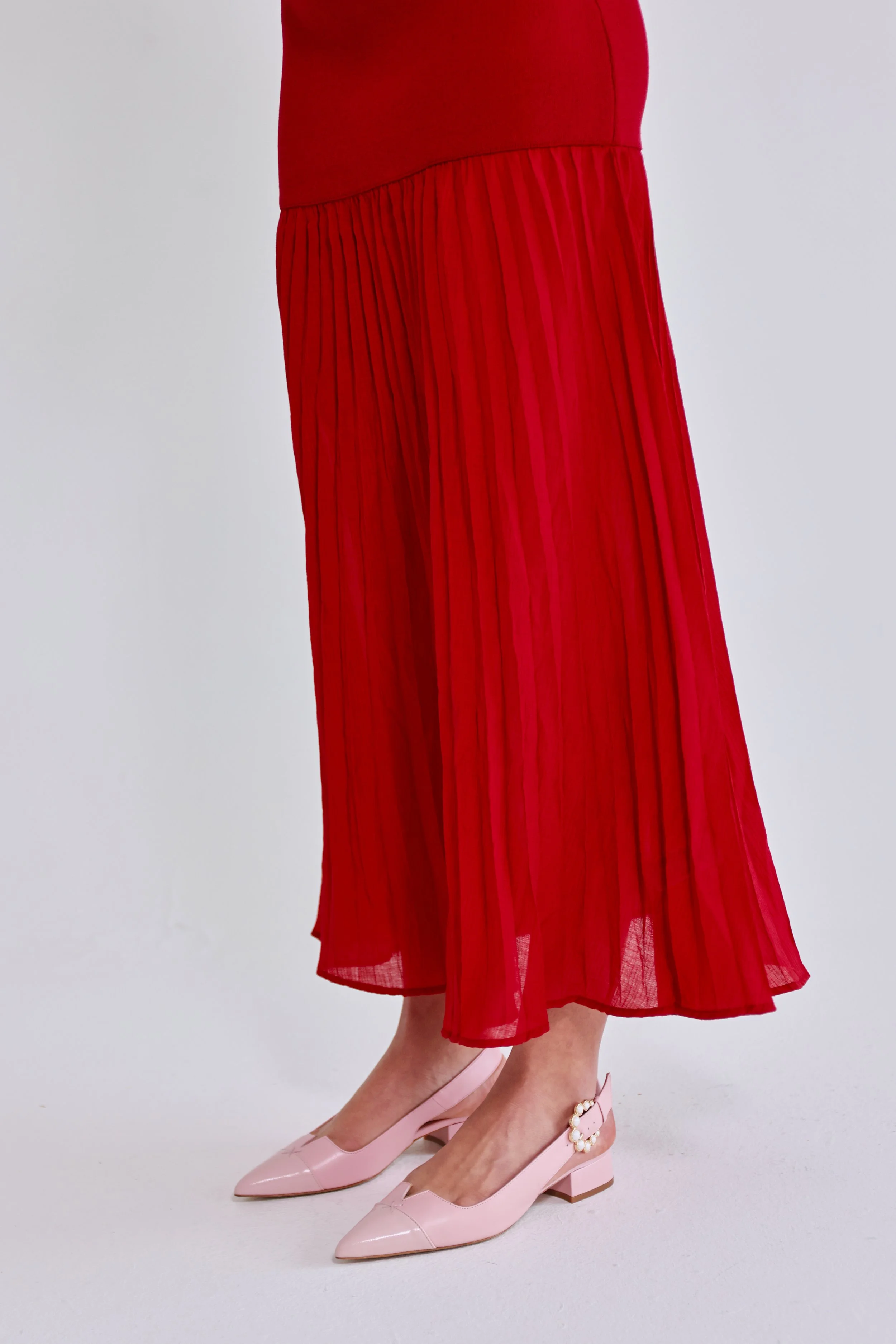 A person wearing a red pleated midi skirt, pink pointed-toe shoes with pearl embellishments on the ankle strap, standing against a plain gray background.