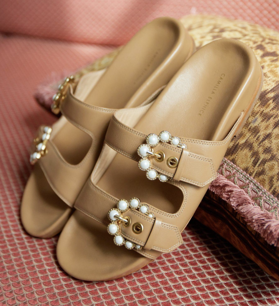 A pair of beige Mary Jane style shoes with pearl embellishments on the straps, placed on a pink textured surface with a patterned cushion nearby.