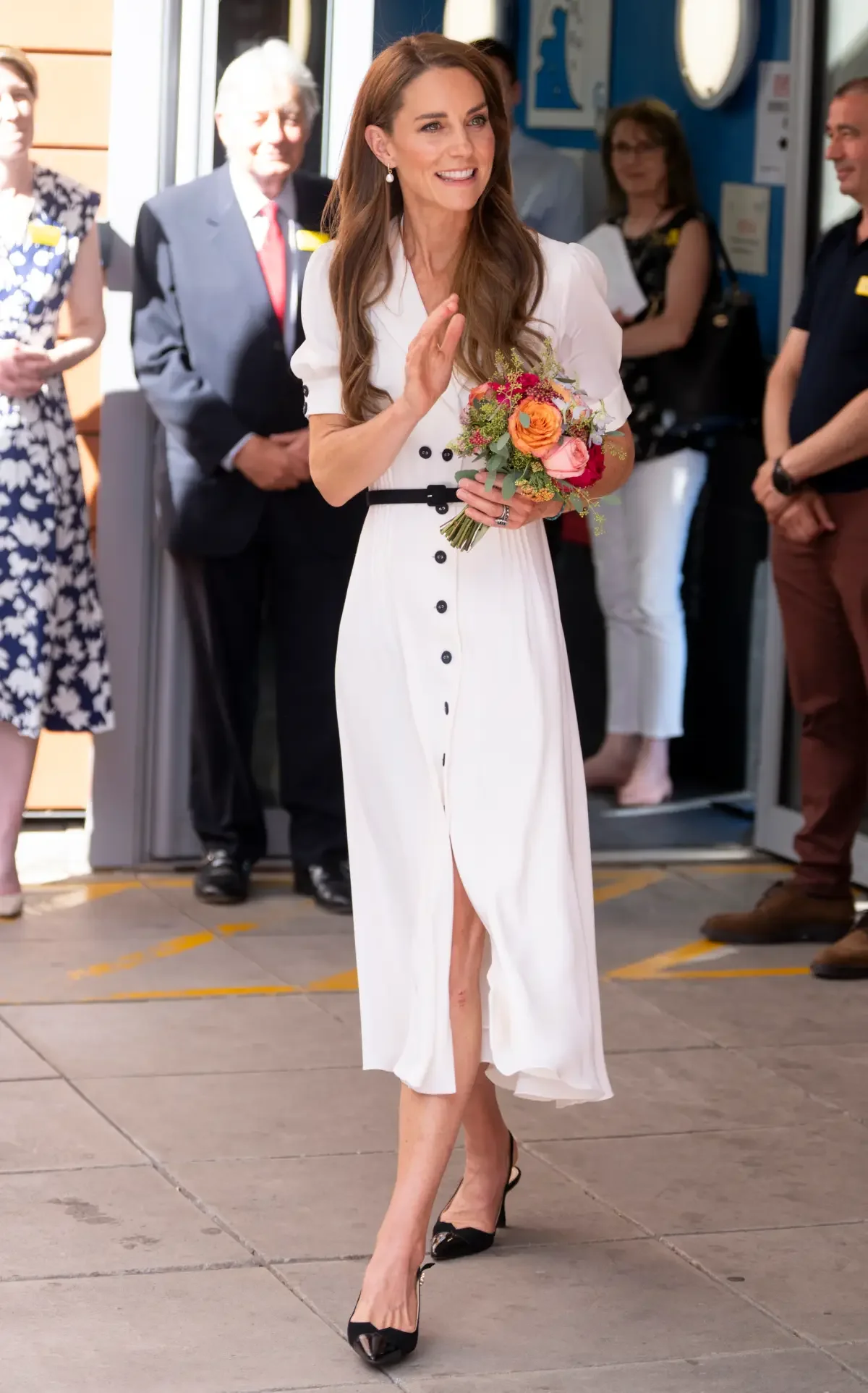 A woman in a white dress holding a colorful bouquet of flowers, smiling as she walks outside in front of onlookers.