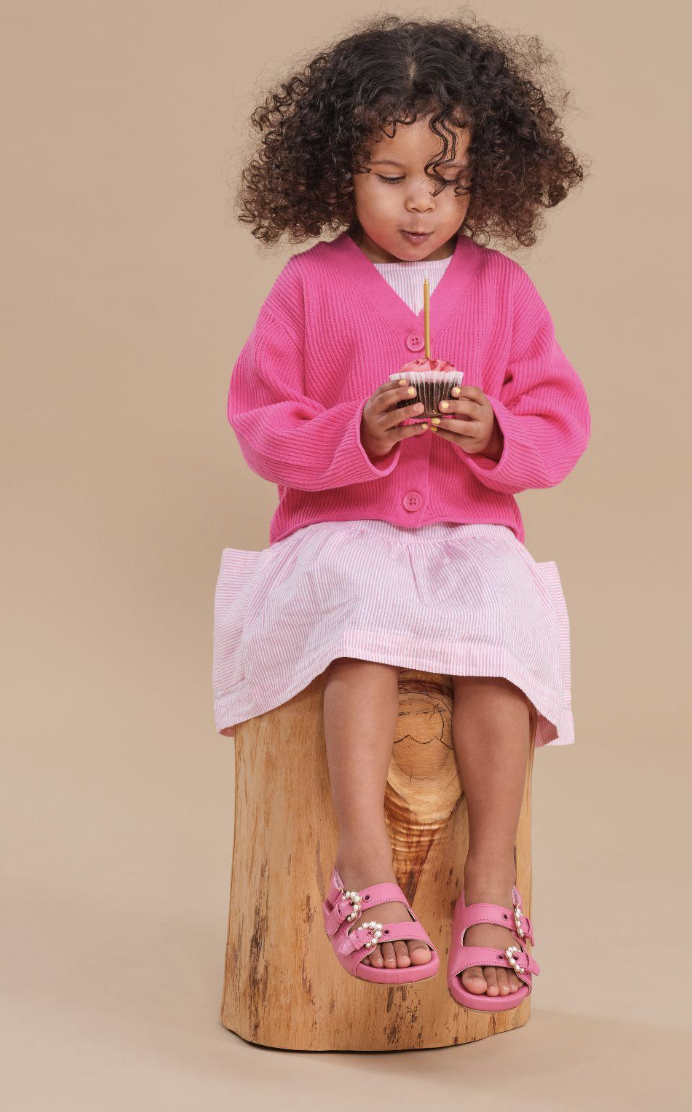 A young girl with curly hair sitting on a wooden stump, holding a cupcake with a candle, wearing a pink cardigan, pink skirt, and pink shoes.