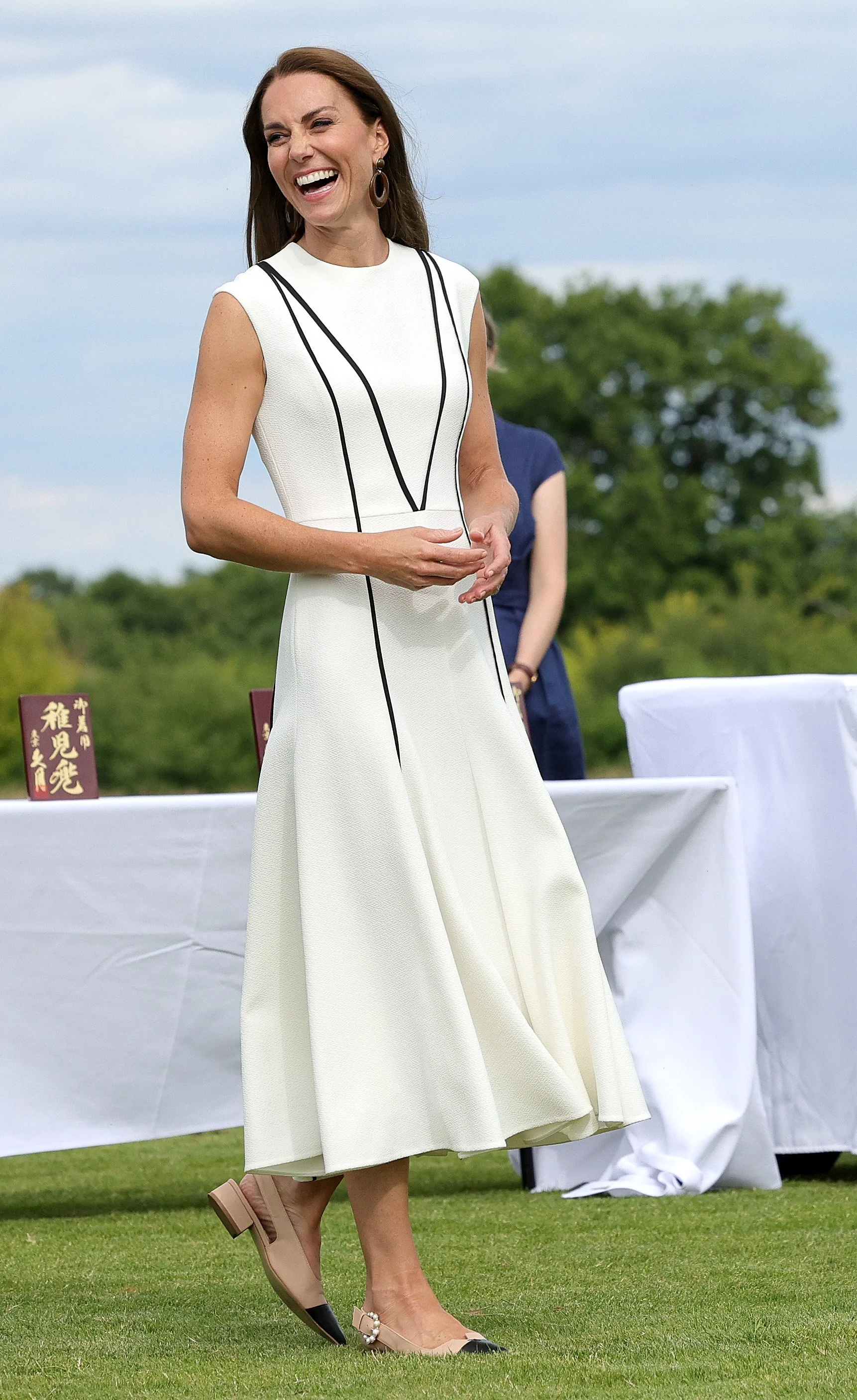 Woman in a sleeveless white dress with black piping, beige heels with black tips and pearl embellishments, standing on grass and smiling outdoors. There is a table with a white cloth and some greenery in the background.