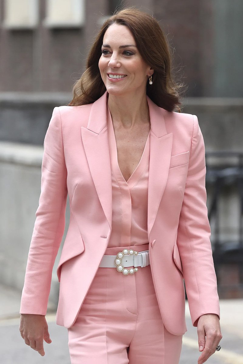A woman wearing a pink suit and a pearl belt, smiling and walking outside in an urban setting.
