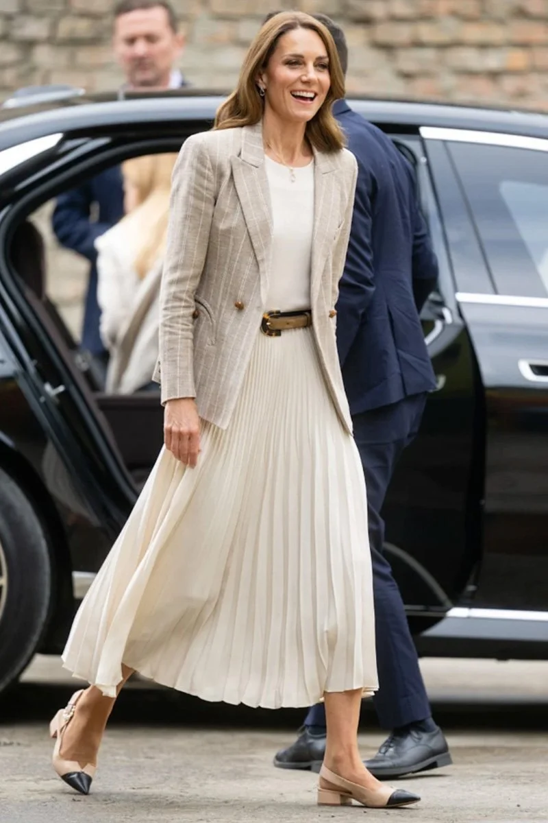 A woman with shoulder-length light brown hair smiling while walking, wearing a beige plaid blazer over a white blouse and a long pleated cream skirt with matching beige and black shoes.
