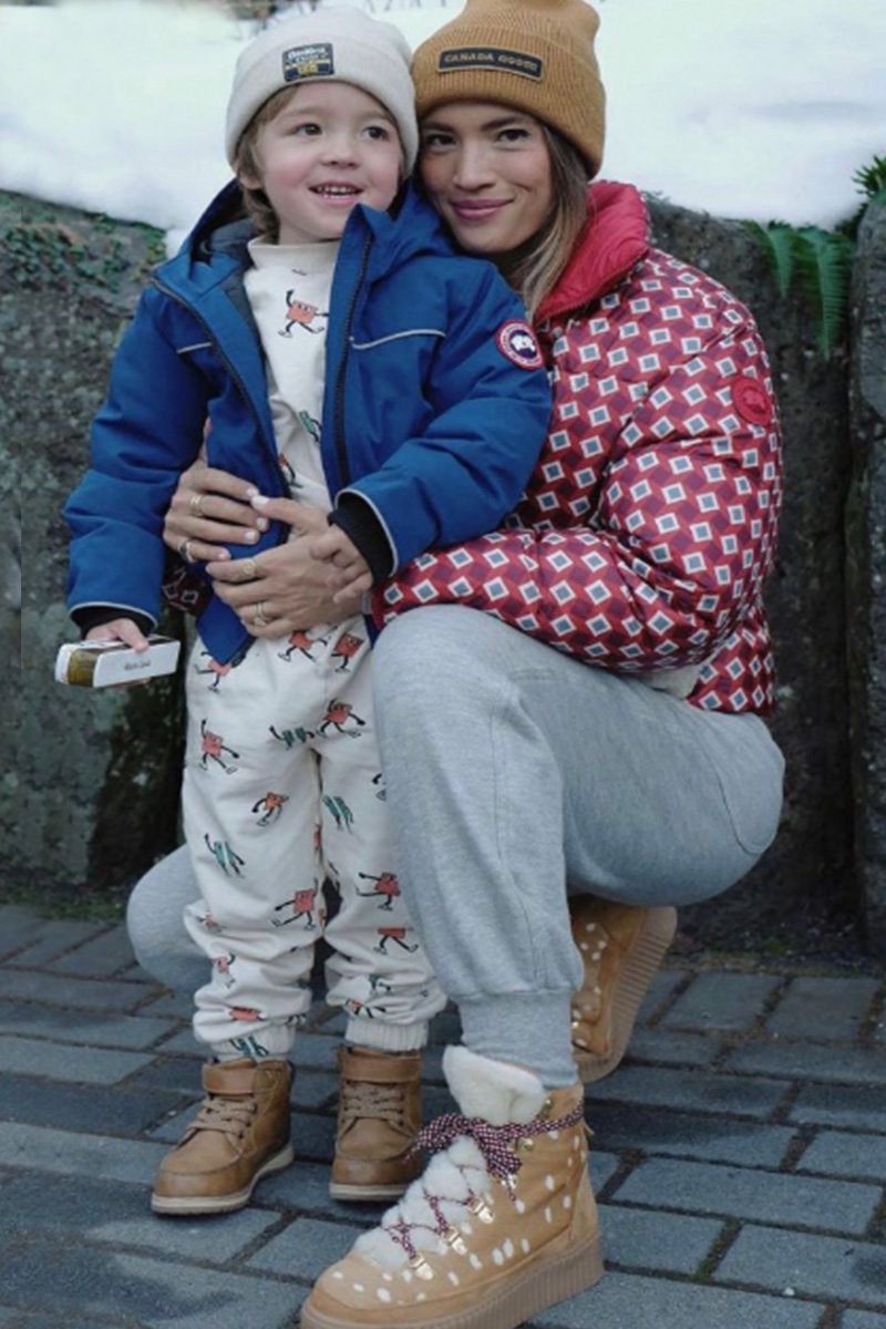 A woman and a young boy are sitting outdoors on a stone surface, dressed in winter clothing, smiling and hugging each other. The woman is wearing a red patterned jacket, beige boots, and gray sweatpants, while the boy is wearing a blue jacket, white 