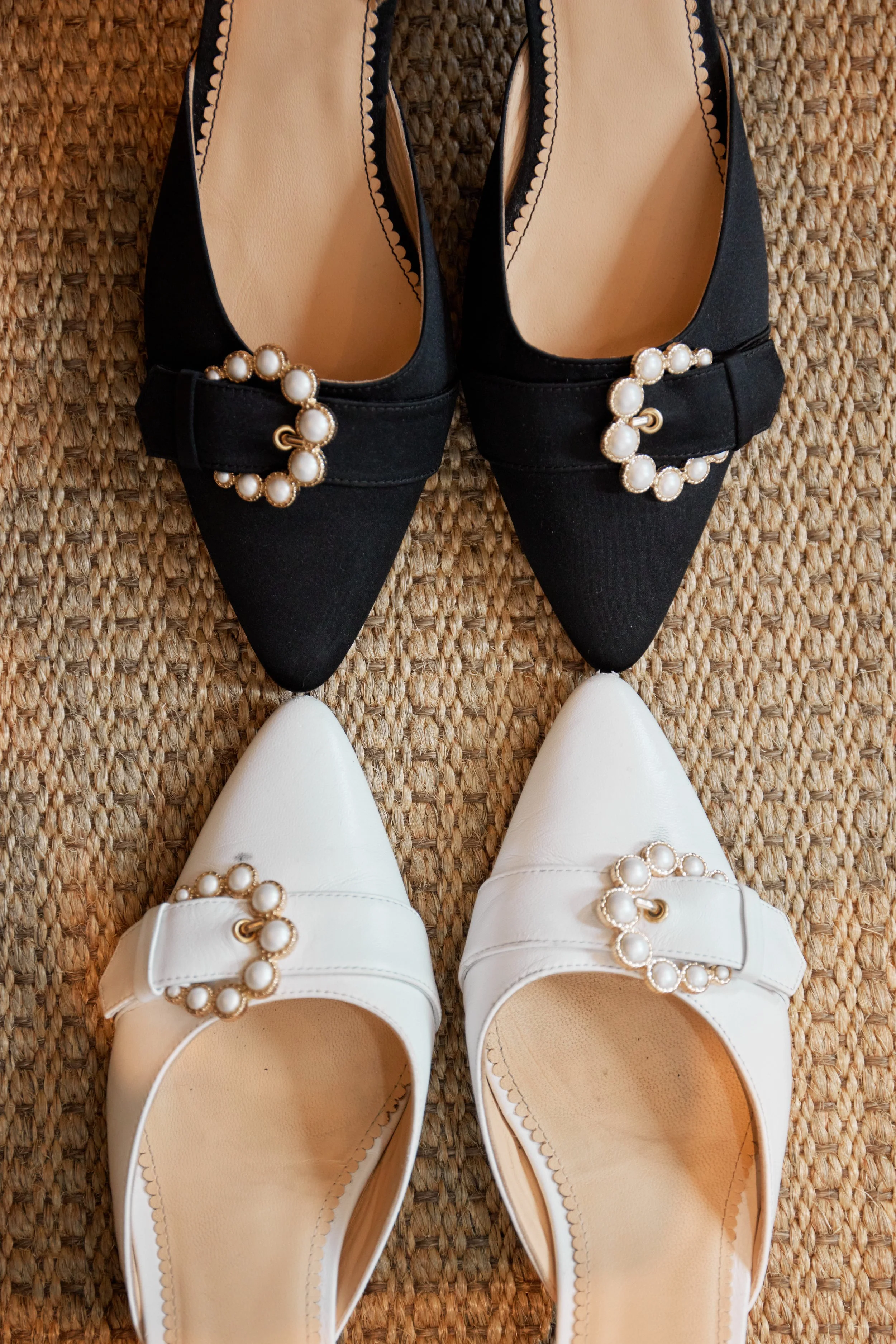 A pair of black pointed-toe shoes with pearl embellishments on a woven rug, and a pair of white pointed-toe shoes with similar pearl decorations, viewed from above.