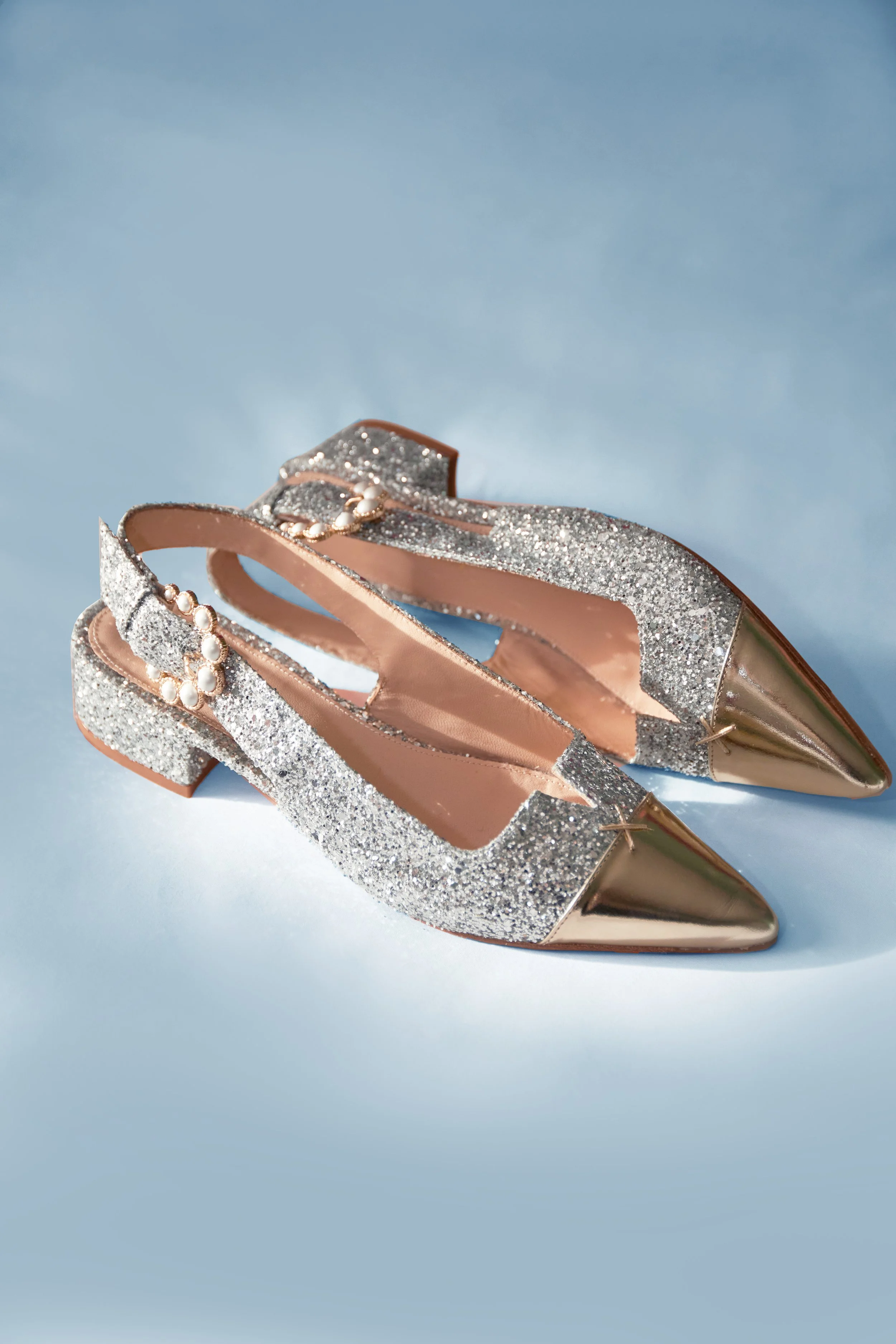 Close-up of a pair of high-heeled shoes with glittery silver straps, pearl accents, and pointed metallic gold toes, set against a light blue background.