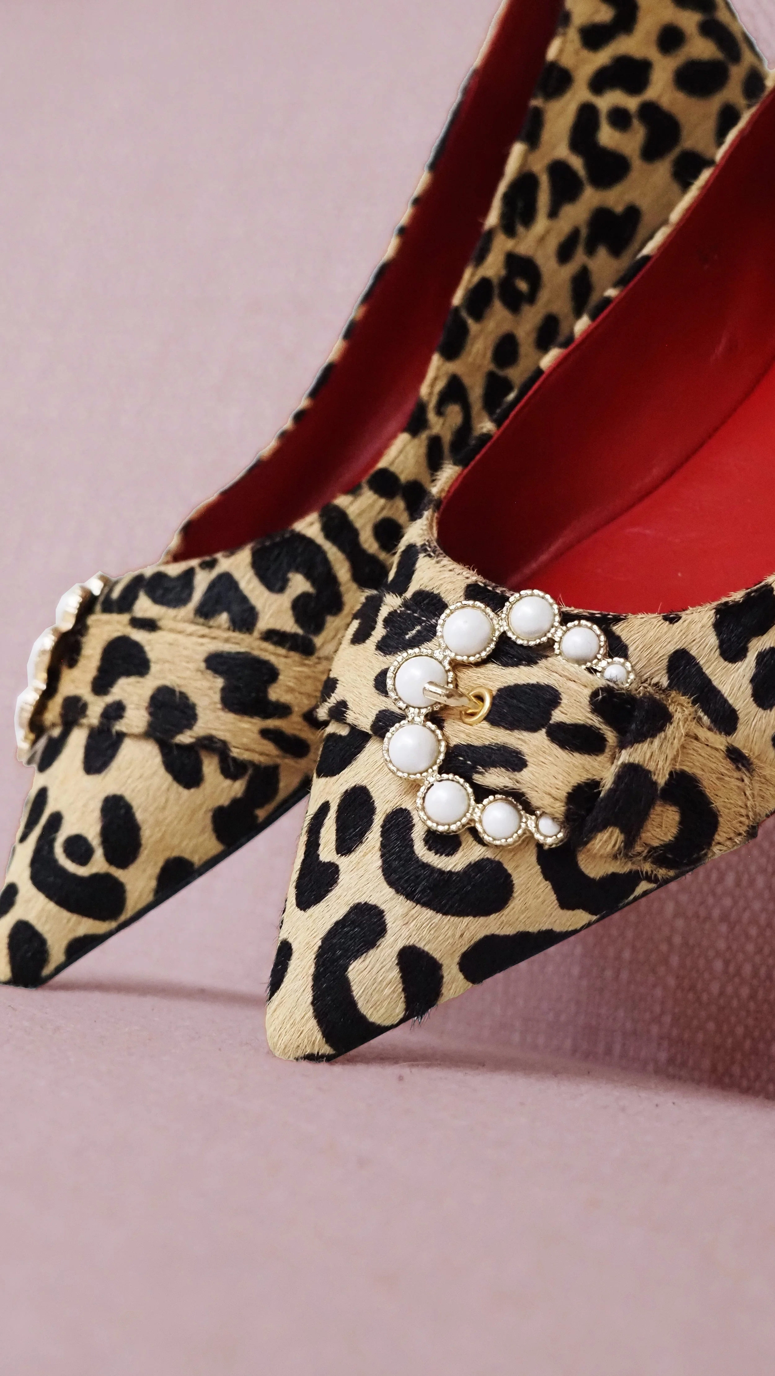 Close-up of a high-heeled shoe with a leopard print pattern, red interior lining, and a pearl and gold embellishment on the front, against a pink background.