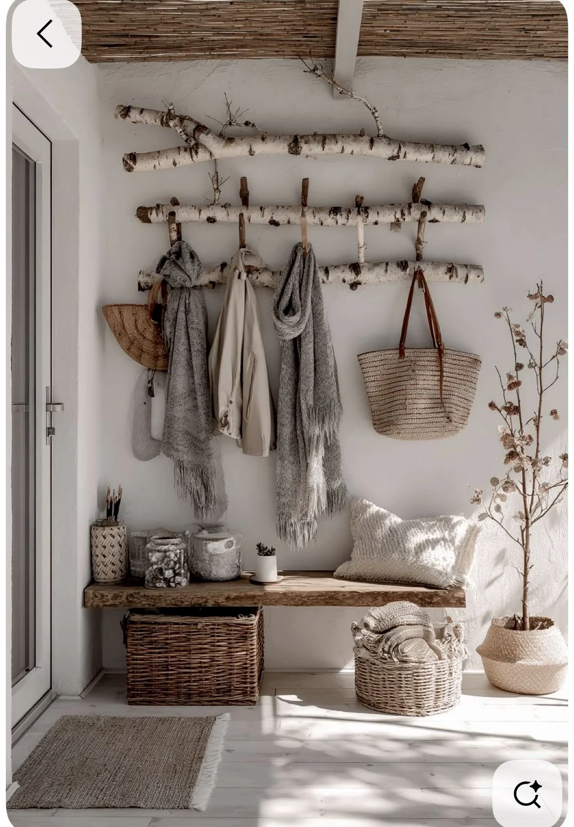 Rustic interior with white walls, birch wood coat rack holding scarves, bags, and jackets, wooden bench with storage baskets, a small table with decorative jars and a potted plant, and a woven pillow on the bench, next to a dried plant in a clay pot.